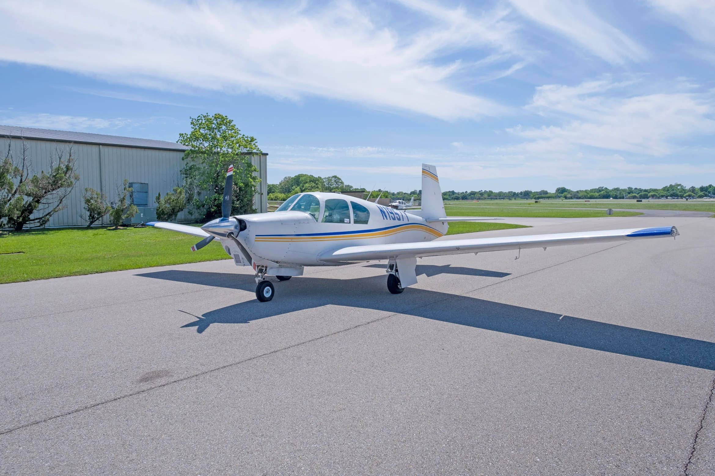A white single-engine propeller aircraft with blue and yellow stripes, registration N1957Y, parked on an asphalt apron next to a hangar.
