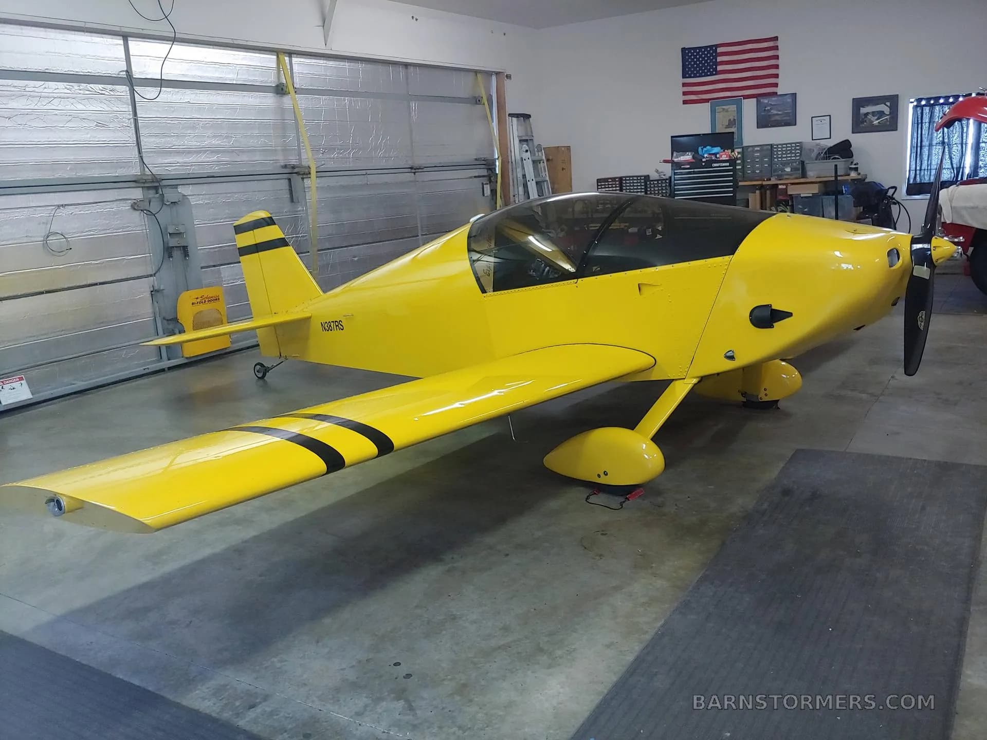 A yellow single-engine aircraft with black stripes on the wing and tail, parked in a garage with an American flag on the wall.