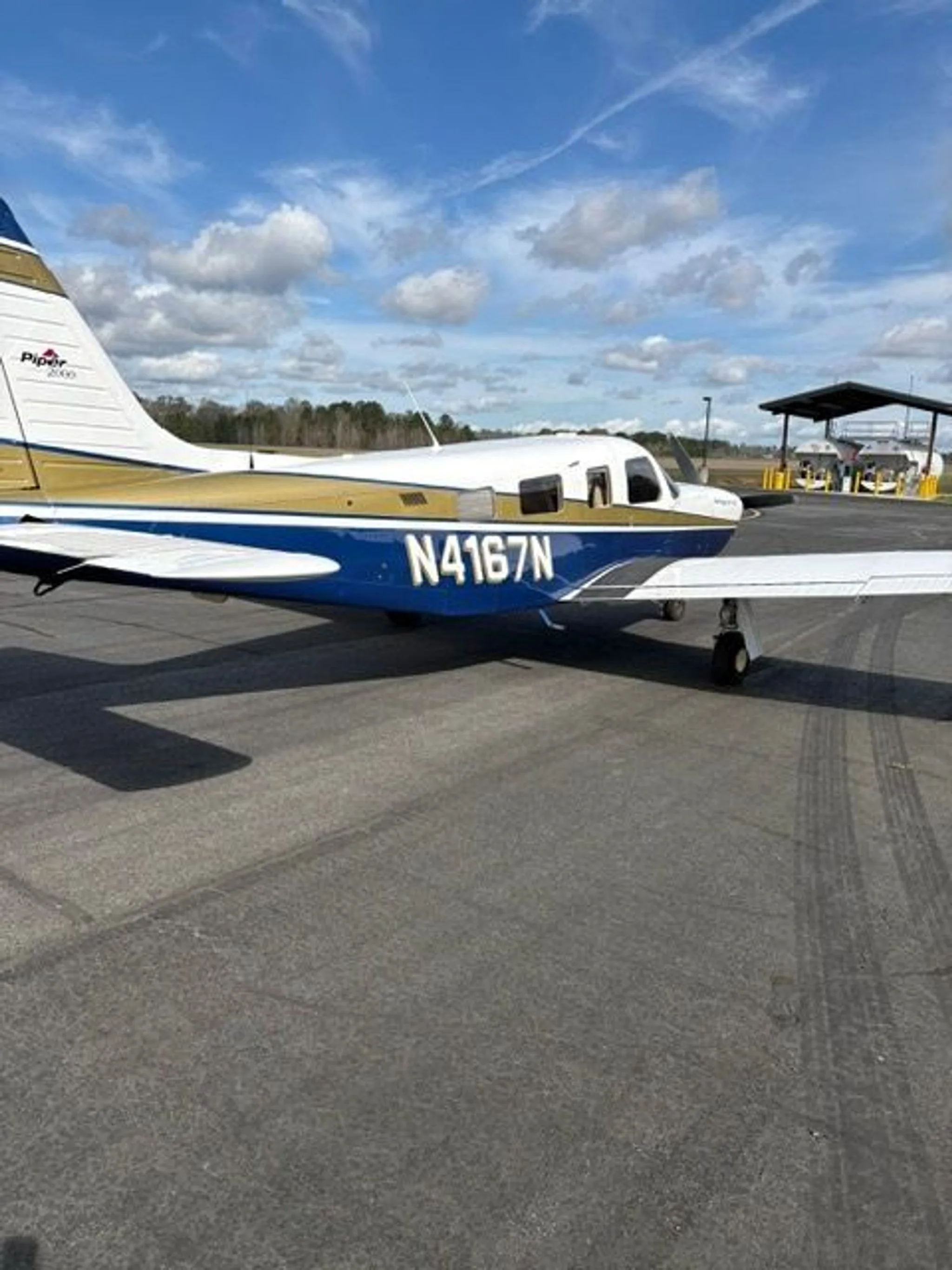 A three-quarter rear view of the Piper Saratoga II TC aircraft, N4167N, with white, blue, and gold paint, parked on an asphalt ramp.