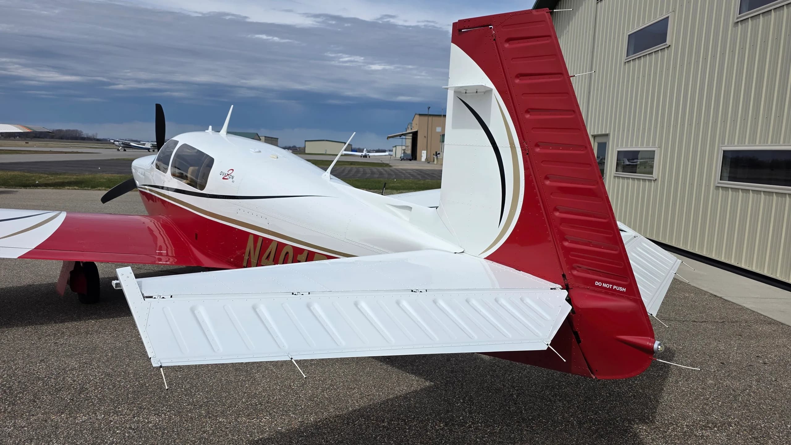 A rear right view of a red and white Mooney aircraft with registration N401PS parked on an asphalt apron next to a hangar.