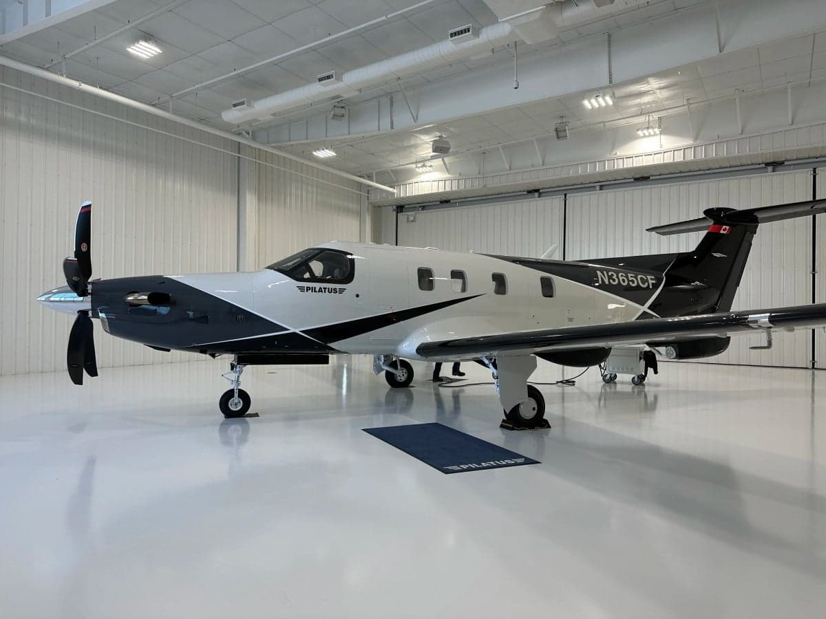 A Pilatus PC-12 aircraft with a gray and white paint scheme is parked inside a hangar.