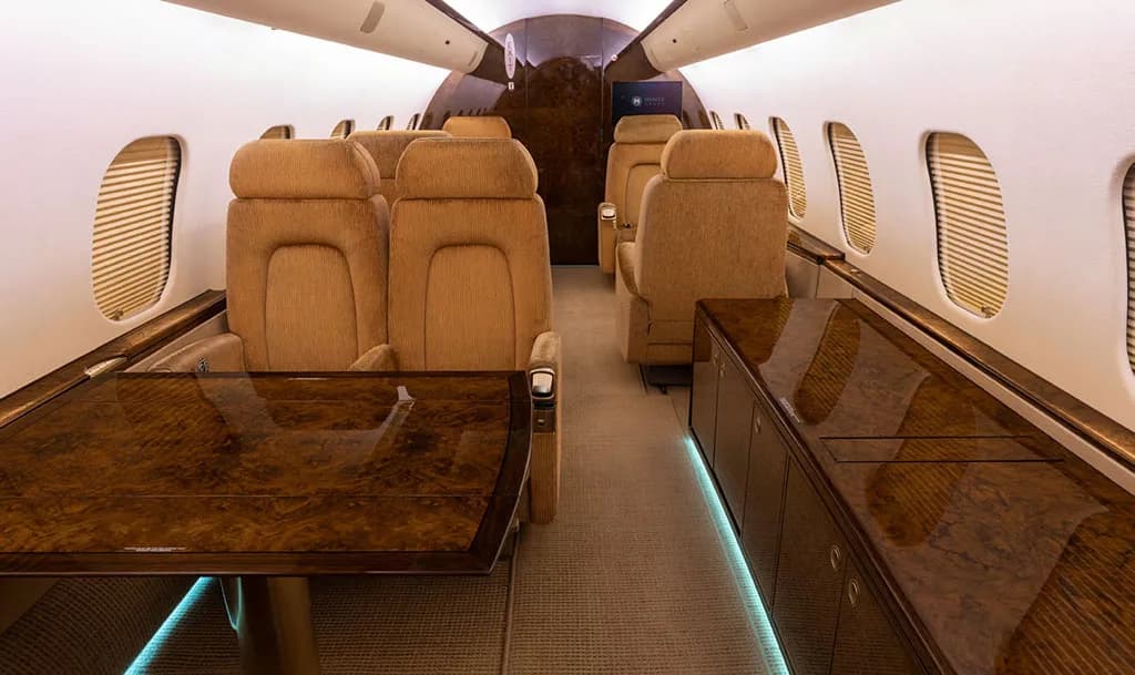 The luxurious interior cabin of a private jet, featuring plush tan seats, a polished dark wood table, and matching cabinetry.