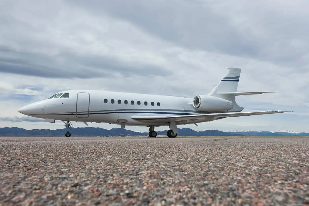 A white Dassault Falcon 2000 business jet with blue stripes on an airfield under a cloudy sky.