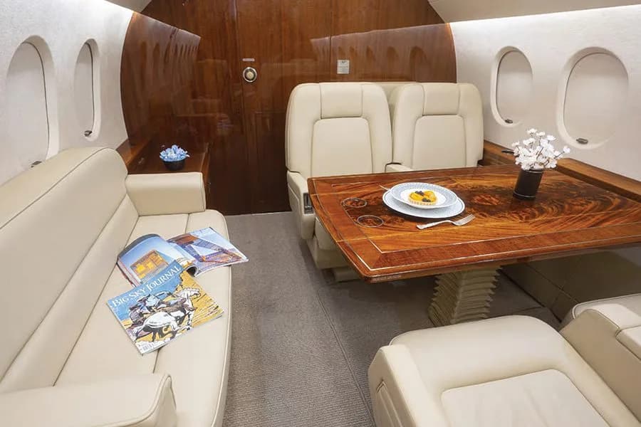 A section of a private jet cabin with cream-colored seating, a polished wooden table set with a plate, and magazines on a side sofa.