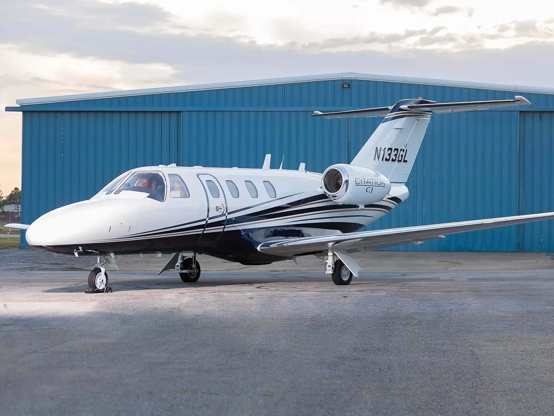 A white Cessna Citation CJ business jet with black and gold stripes is parked on an asphalt ramp in front of a blue hangar.