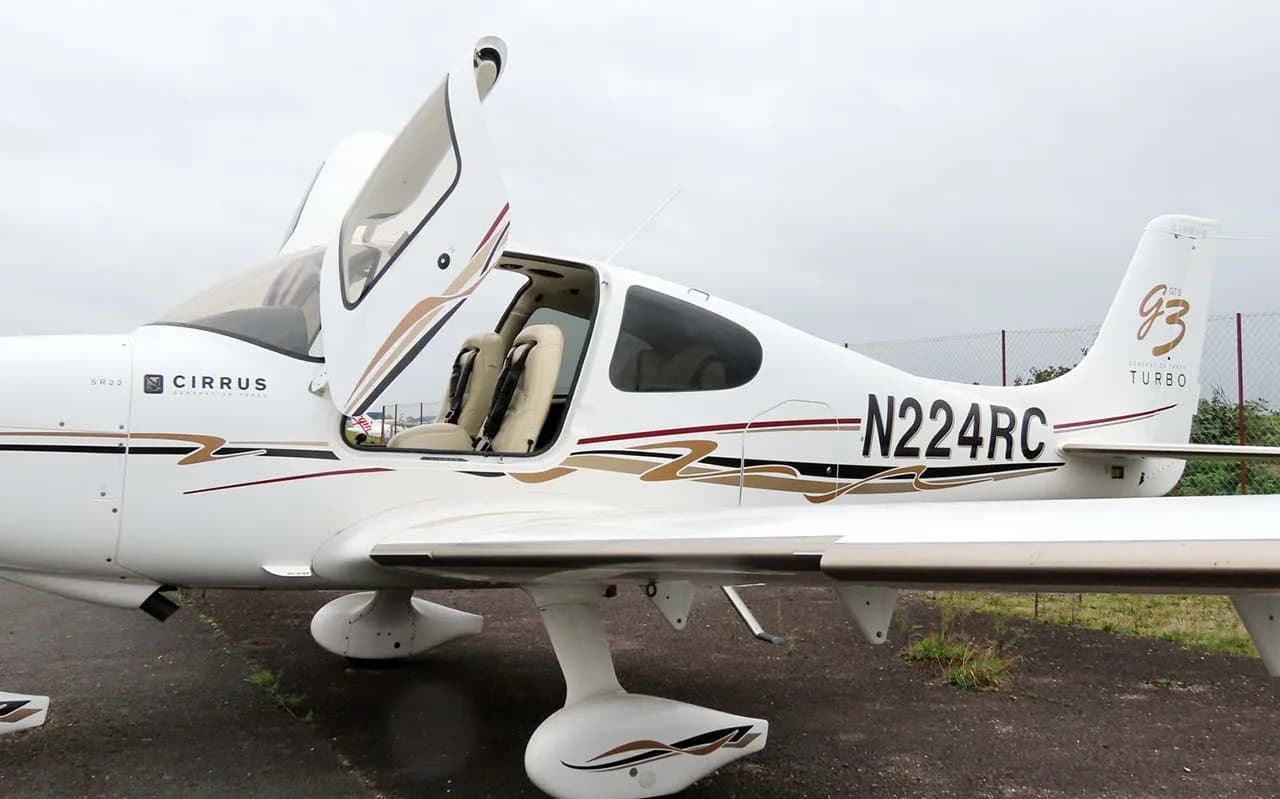 A side view of a white Cirrus SR22T (N224RC) with its gull-wing doors open, revealing the light-colored interior.
