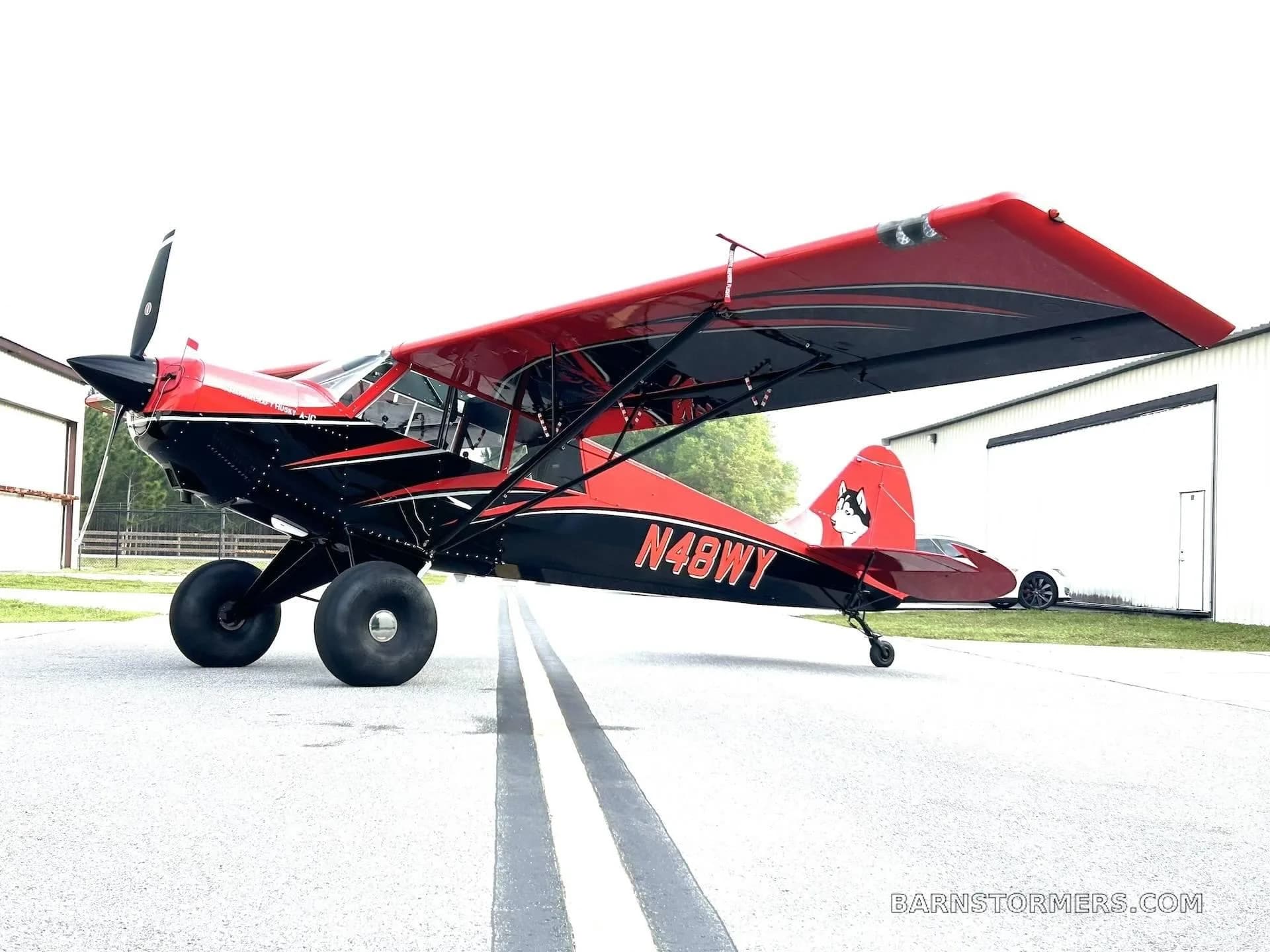 A red and black Aviat Husky A-1C aircraft with large tundra tires is parked on a tarmac in front of a white hangar.