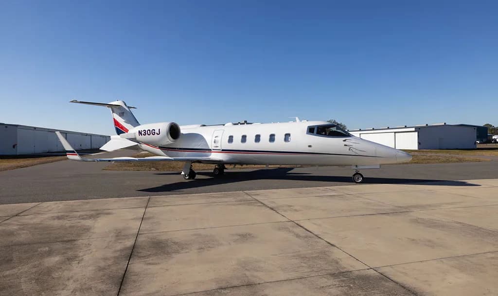 A white Learjet with red and blue tail stripes, registration N30GJ, parked on an airfield under a clear blue sky.