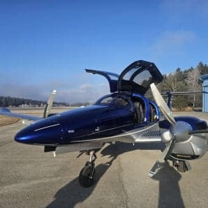 A blue and silver Diamond DA62 aircraft is parked on the tarmac with its gull-wing doors open.