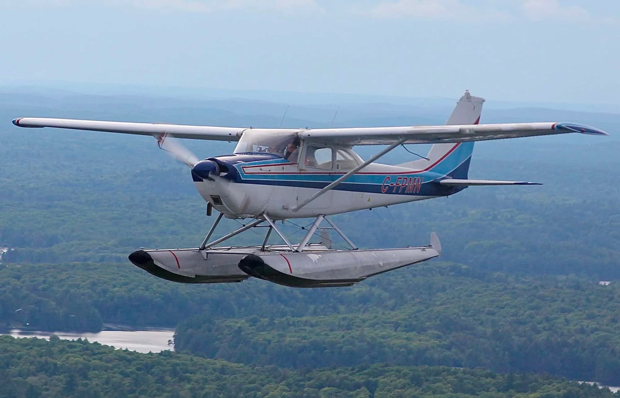 A white and blue Cessna 172 floatplane, registration C-FPMN, flies over a forested landscape with water below.