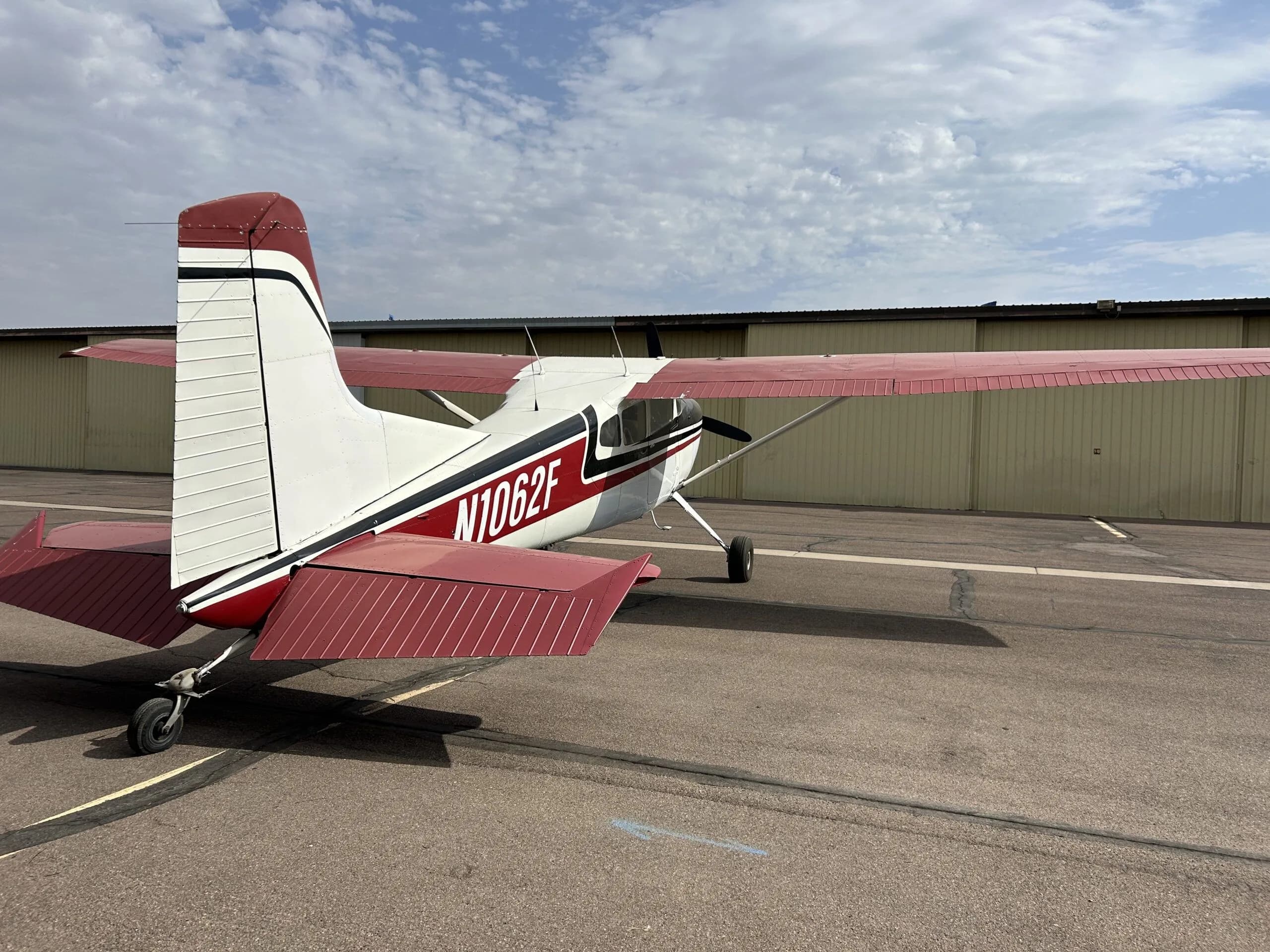 A white, black, and red Cessna 180 aircraft (N1062F) parked on an airport tarmac, viewed from the rear-right showing the tail.