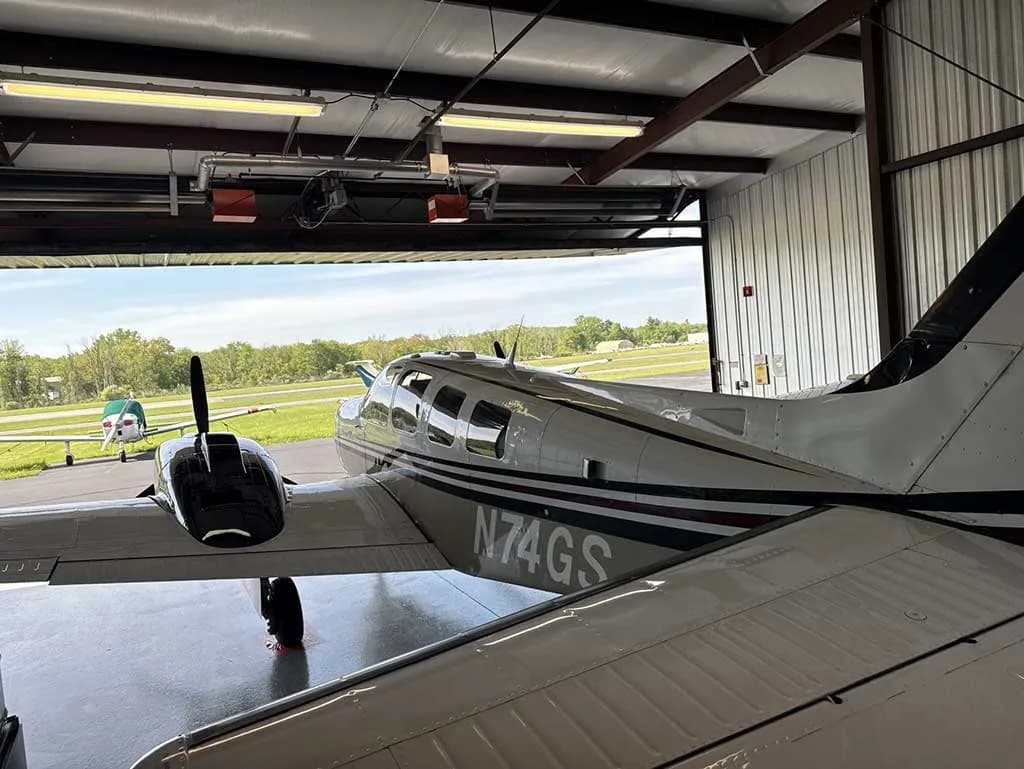A rear view of the aircraft N74GS inside a hangar, looking out onto an airfield with another small plane in the distance.