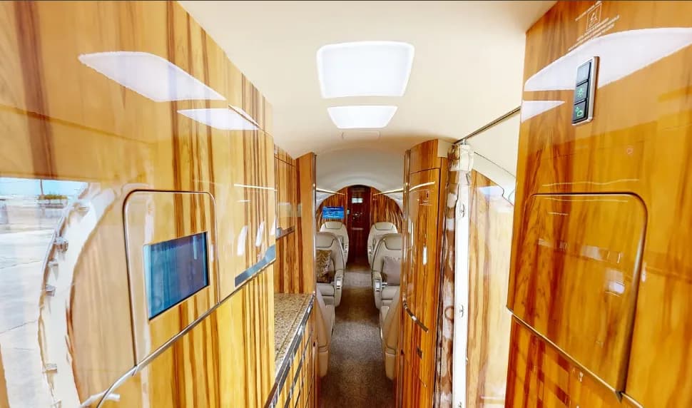 A view down the aisle of a private jet cabin, showcasing the galley area with polished wood and light-colored seating in the background.
