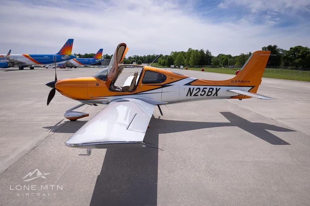 A rear-right view of the orange and silver Cirrus SR22T N25BX with its doors open on an airfield.