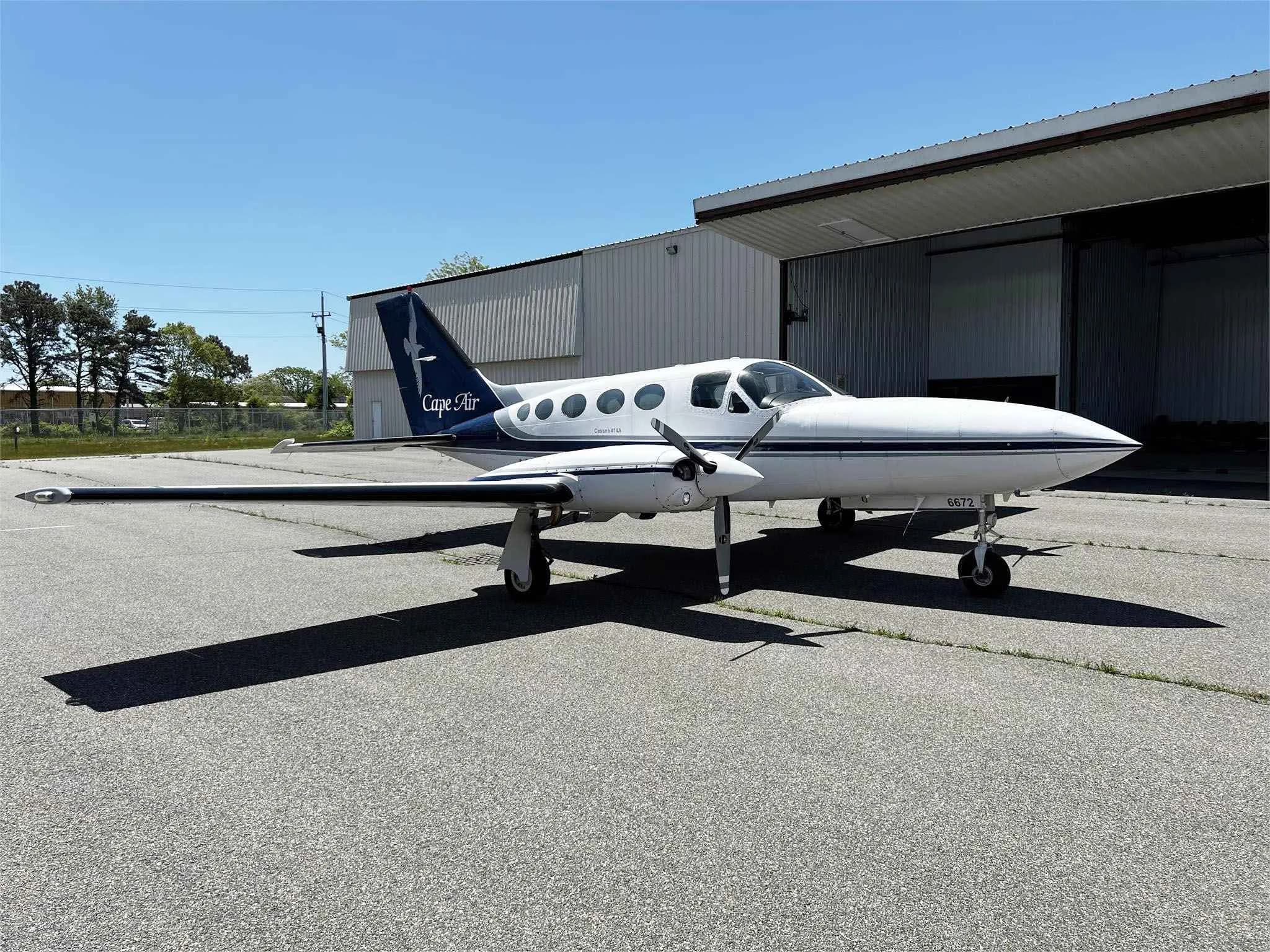 A white and blue Cessna 414A with 'Cape Air' on its tail is parked on an asphalt tarmac next to an open hangar on a sunny day.