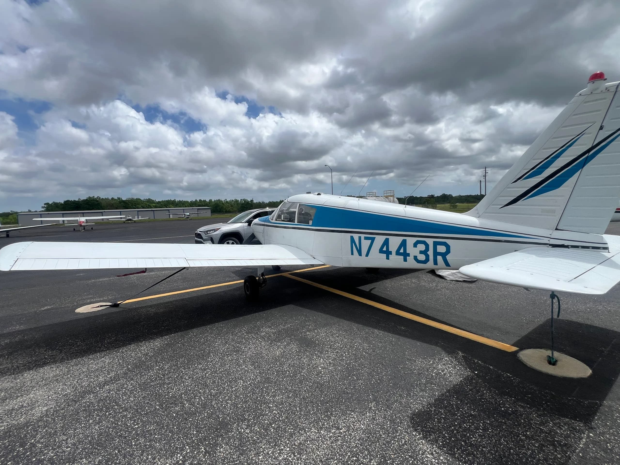 Rear-side view of a white and blue Piper Cherokee 140 (N7443R) parked on an asphalt ramp.