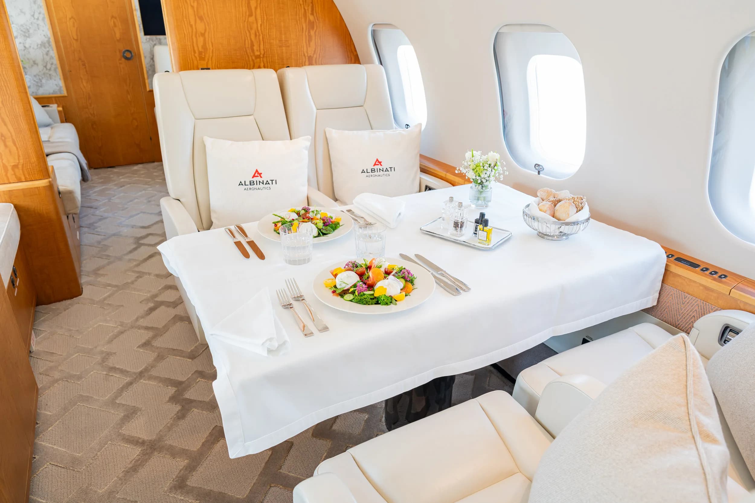 A dining table set with salads and bread inside a private jet cabin, featuring "ALBINATI AERONAUTICS" pillows.