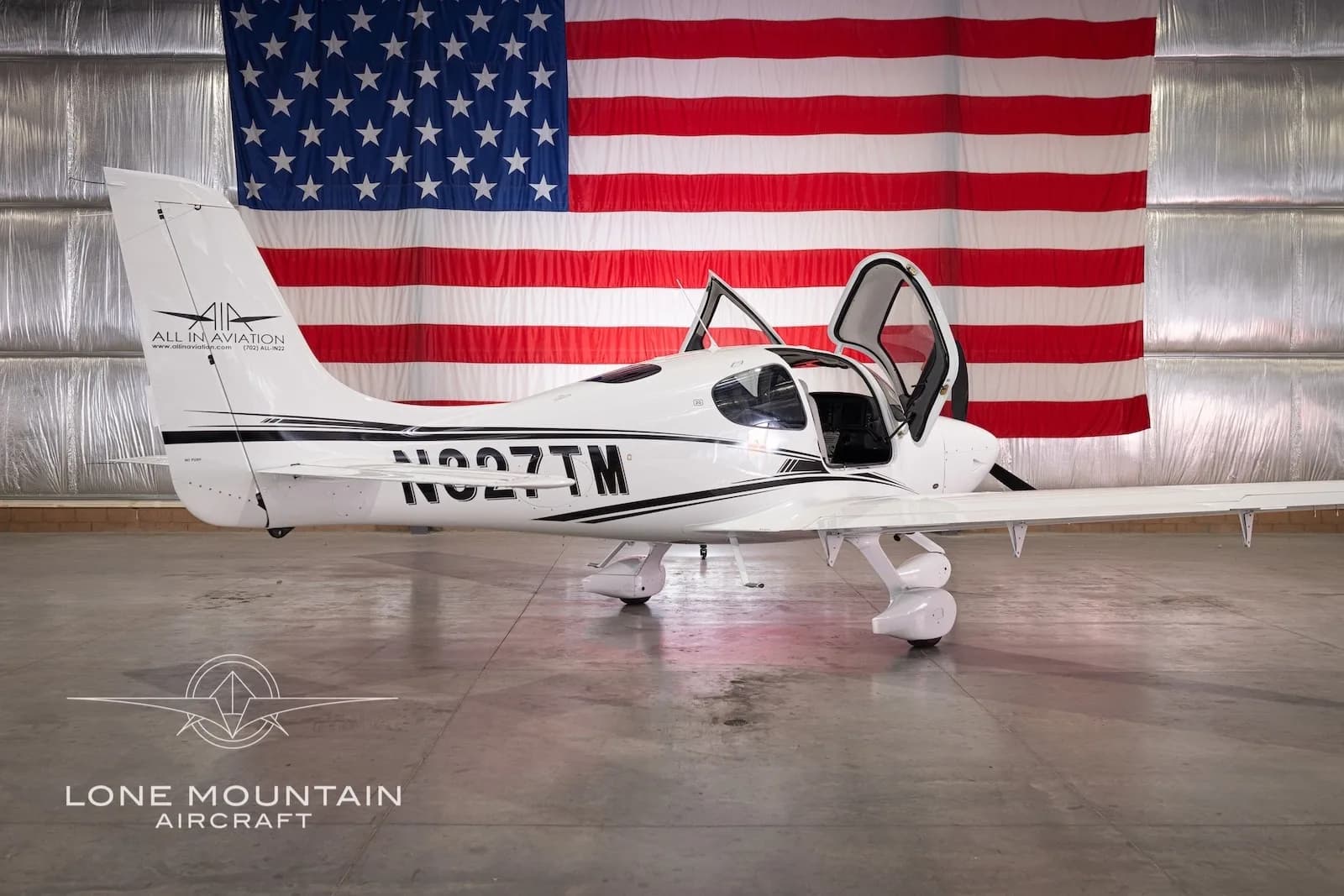 Rear-side view of a white Cirrus SR22 aircraft with black stripes and registration N327TM, parked in a hangar with an American flag backdrop, with its gull-wing doors open.