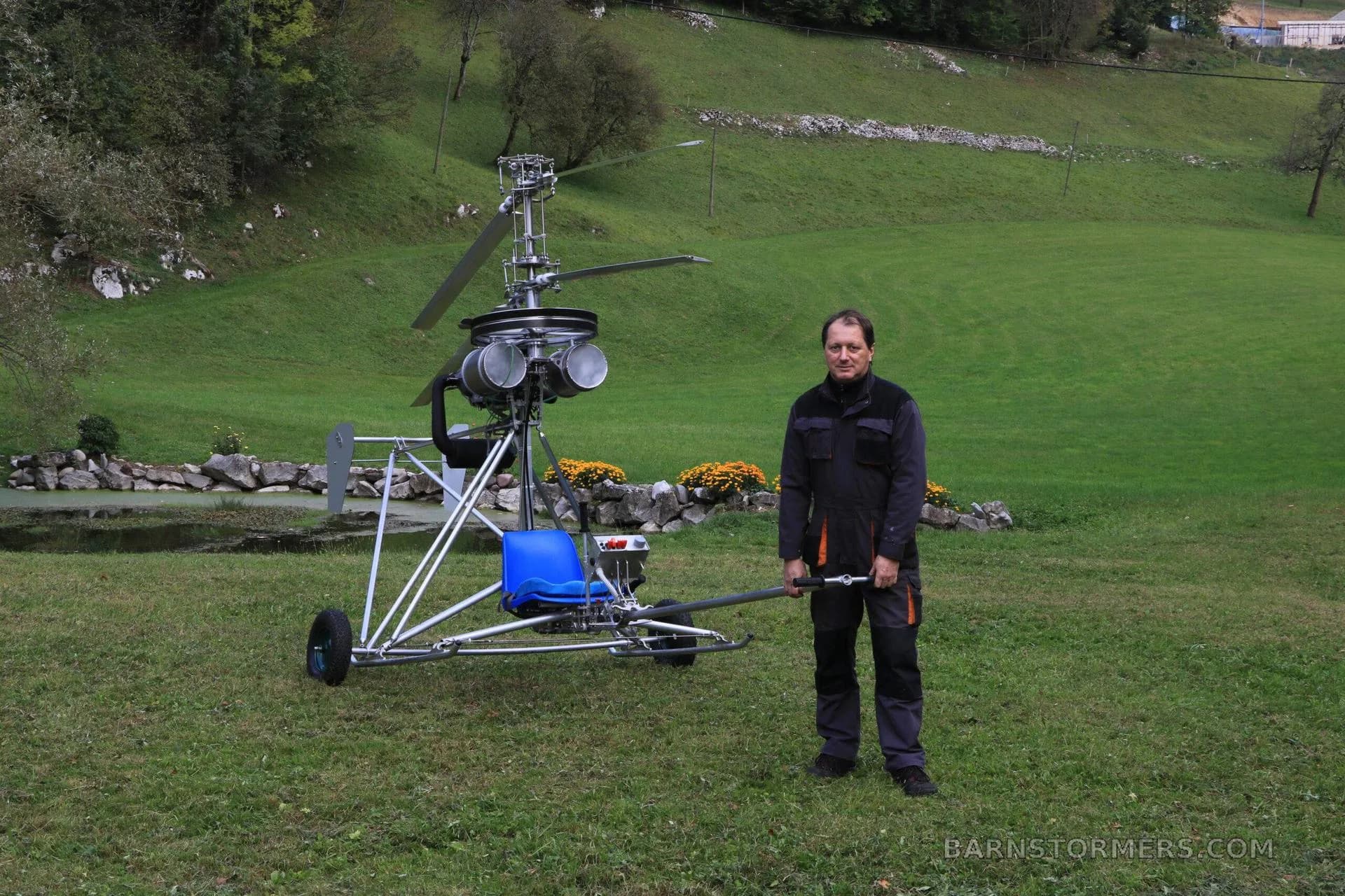 A man in a dark jacket stands next to a single-seat ultralight helicopter with wheels on a green lawn.