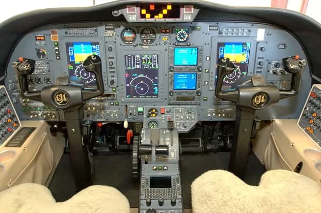 The cockpit of a modern business jet, featuring multiple digital displays, control yokes, and a central throttle quadrant.
