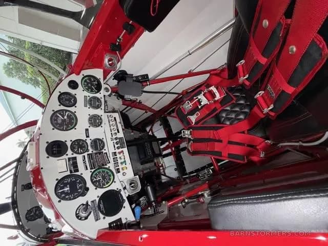 The cockpit of the red biplane, showing the instrument panel with multiple gauges and a red five-point harness on the pilot's seat.