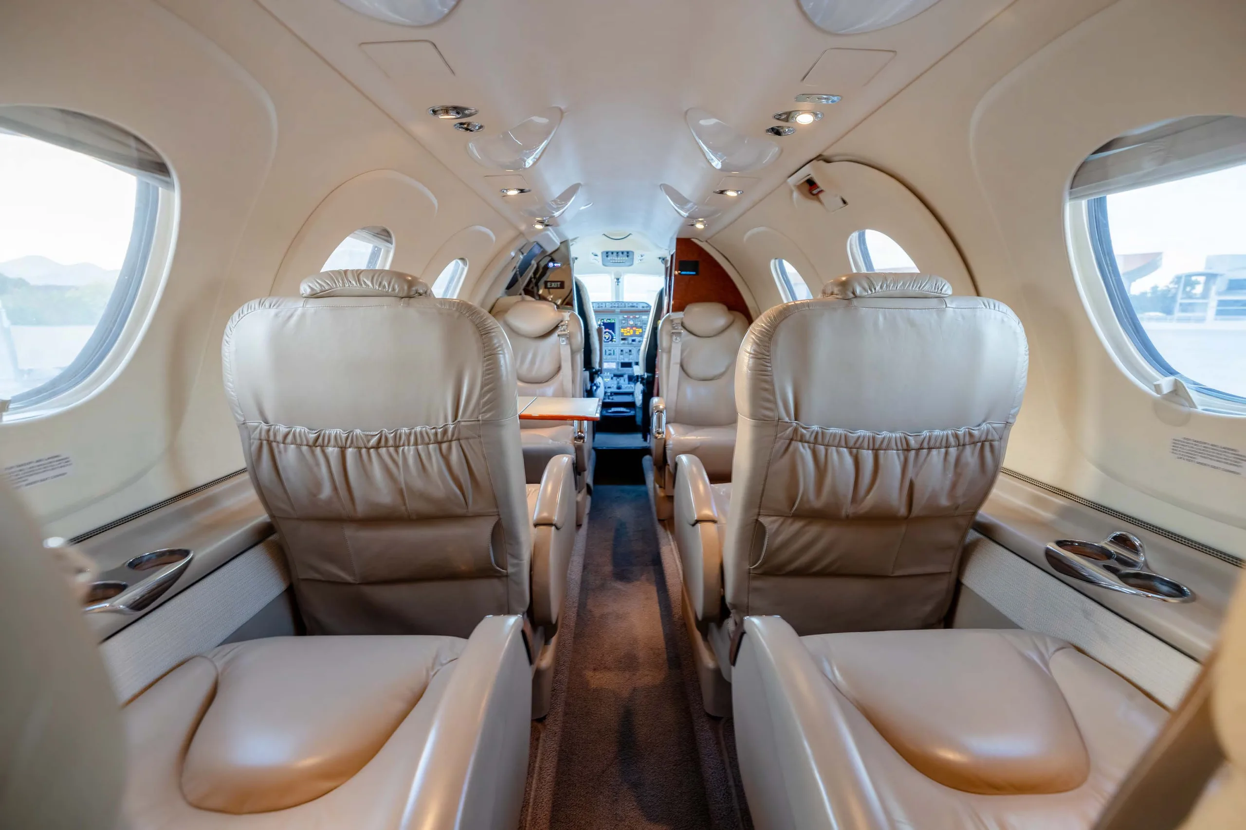 A view down the aisle of a business jet cabin, showing light tan leather seats and the cockpit in the distance.