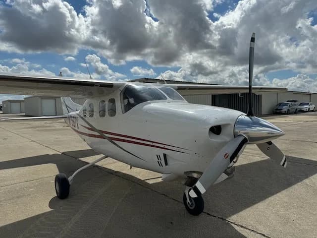 A front view of a white Cessna 206 with red and grey stripes, parked on an airfield with hangars in the background.