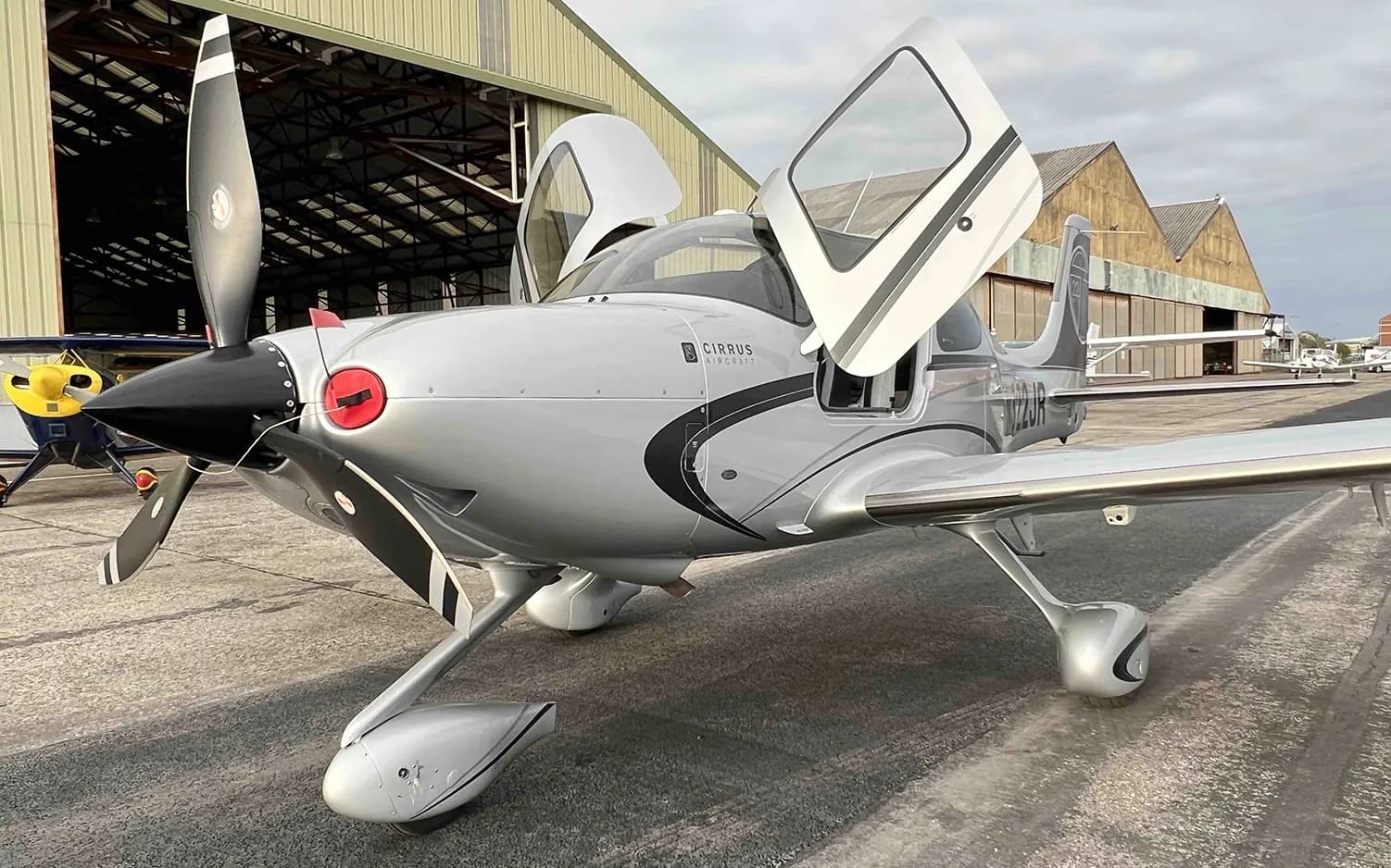 A silver Cirrus SR22 aircraft with black accents and open doors is parked in front of a hangar.
