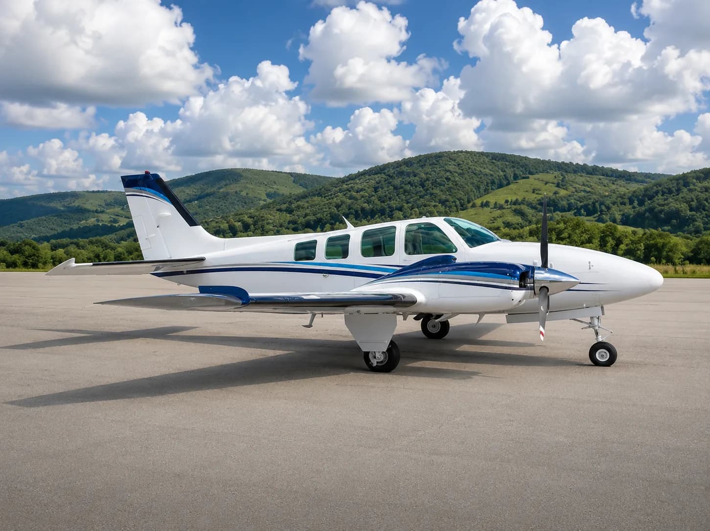 A white and blue twin-engine propeller aircraft is parked on an asphalt runway with green hills and a cloudy sky in the background.