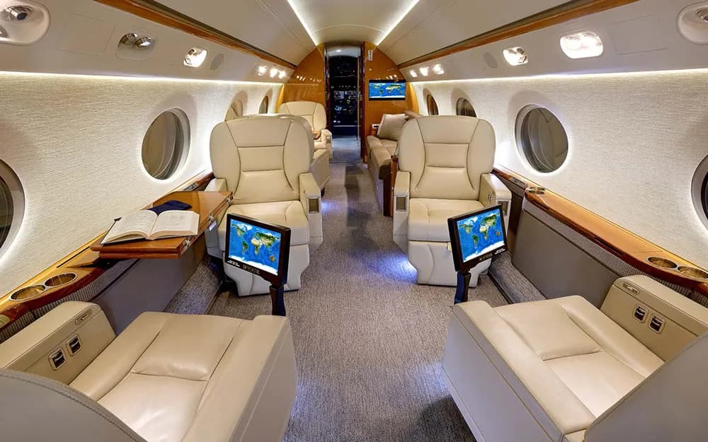 The aircraft cabin with multiple beige seats, individual entertainment screens displaying a world map, and a book on a side table.