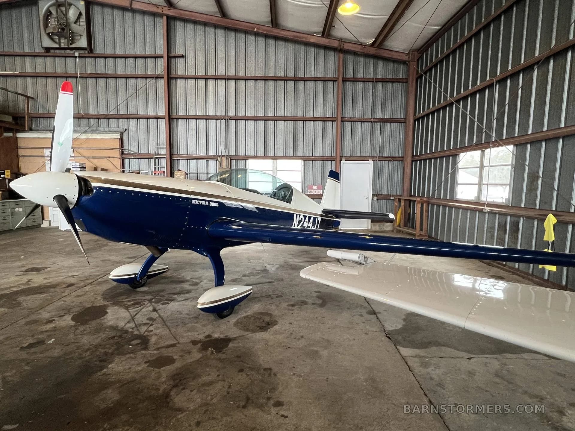 A blue and white Extra 300L aircraft with tail number N244JT is parked inside a metal hangar, viewed from the front-left.