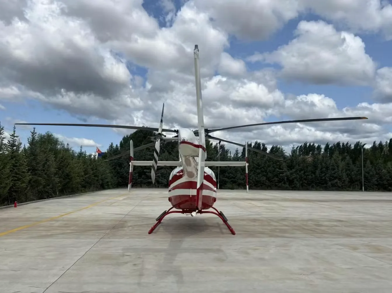 A rear view of the white and red Bell 407 GXi helicopter showing its tail rotor and skids on a concrete pad.