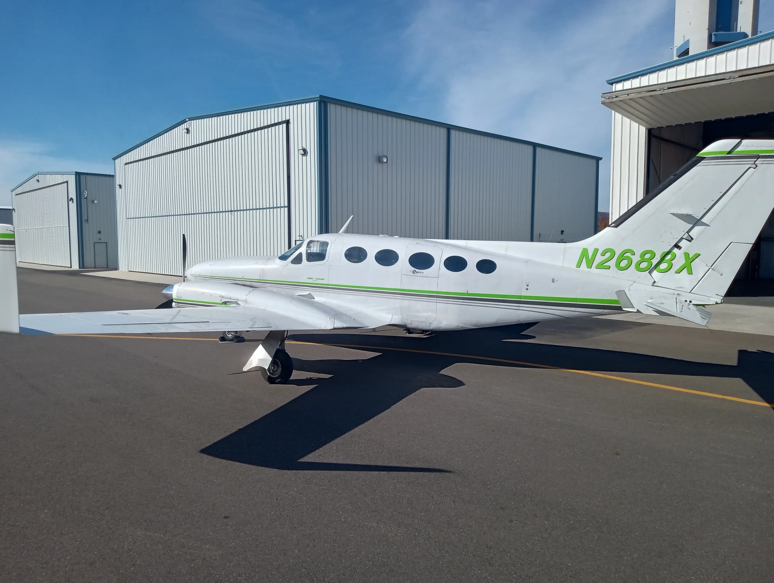 A white twin-engine propeller aircraft with green and black stripes, N2688X on the tail, parked on an asphalt ramp with hangars in the background.