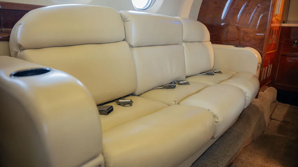 A cream leather three-person sofa seat in the private jet cabin, with cup holders and seatbelts visible.