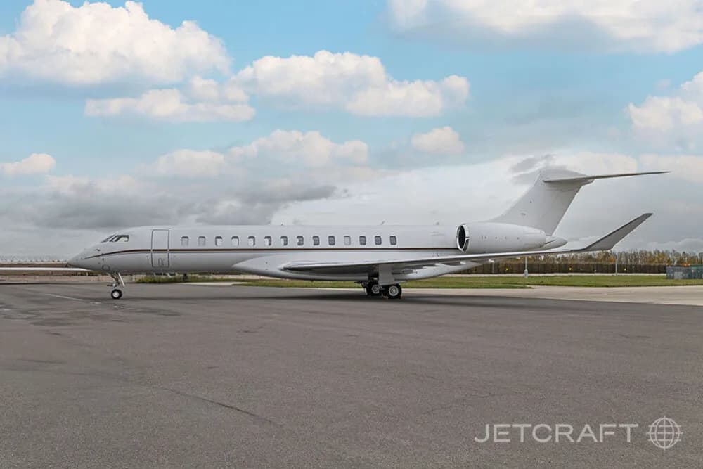 A full side view of a white Bombardier Global business jet with brown accent stripes parked on an airfield.