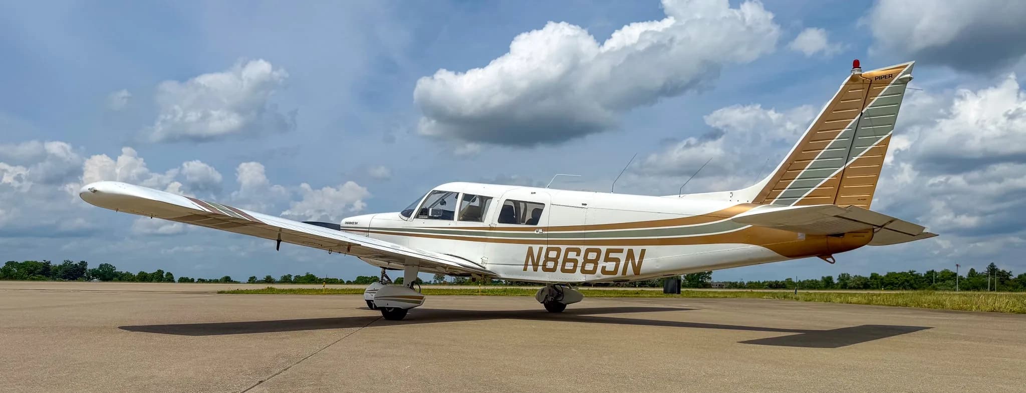 A rear-left view of a white Piper Cherokee Six aircraft with brown and green stripes, showing registration N8685N, parked on an airfield.