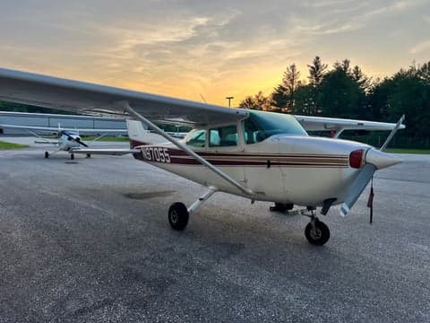 A white Cessna 172, N97055, with maroon and gold stripes, parked on an airfield at sunset with another aircraft in the background.