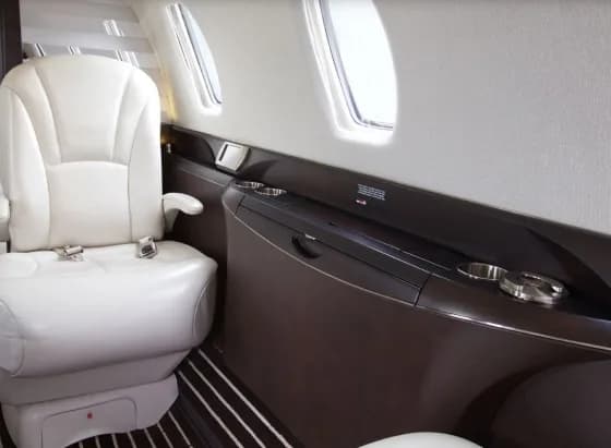 A close-up shows a white leather cabin seat next to a dark wood panel with cup holders and a storage compartment.