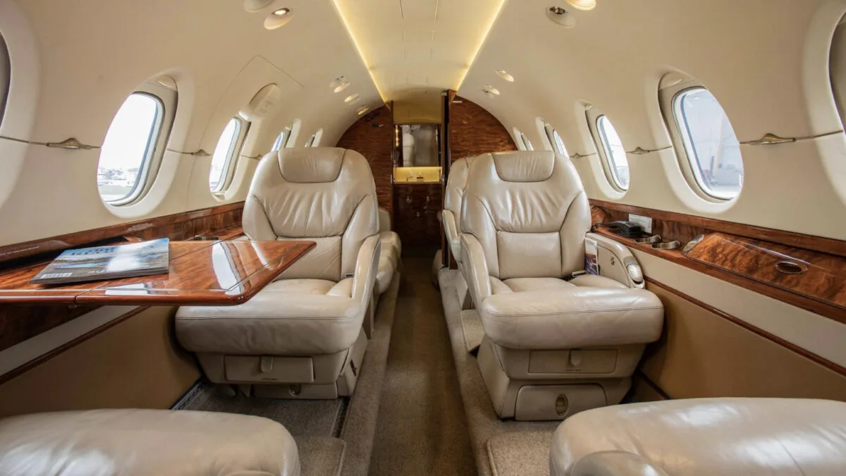 The luxurious interior cabin of a business jet with beige leather seats, wood trim, and a small table.