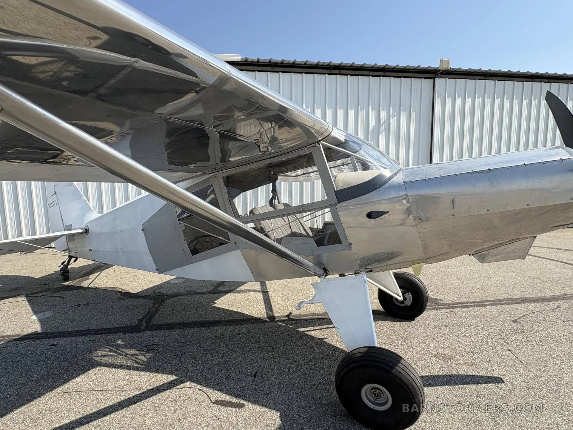 A side view of a silver and white single-engine aircraft, registration N213TJ, parked on an asphalt tarmac next to a white hangar.