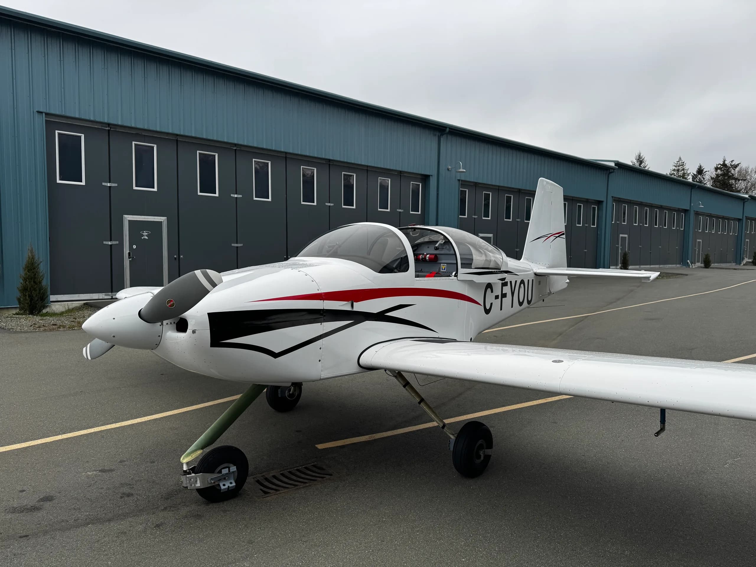 A white single-engine aircraft, registration C-FYOU, with red and black stripes, parked on an asphalt apron in front of blue hangars.