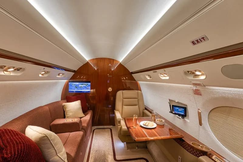 A section of a business jet's cabin interior featuring a brown sofa, a single tan seat, and a wooden table with snacks.