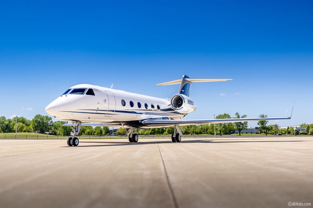The N999GC Gulfstream G550 business jet, white with blue and black stripes, parked on an airfield under a clear blue sky.