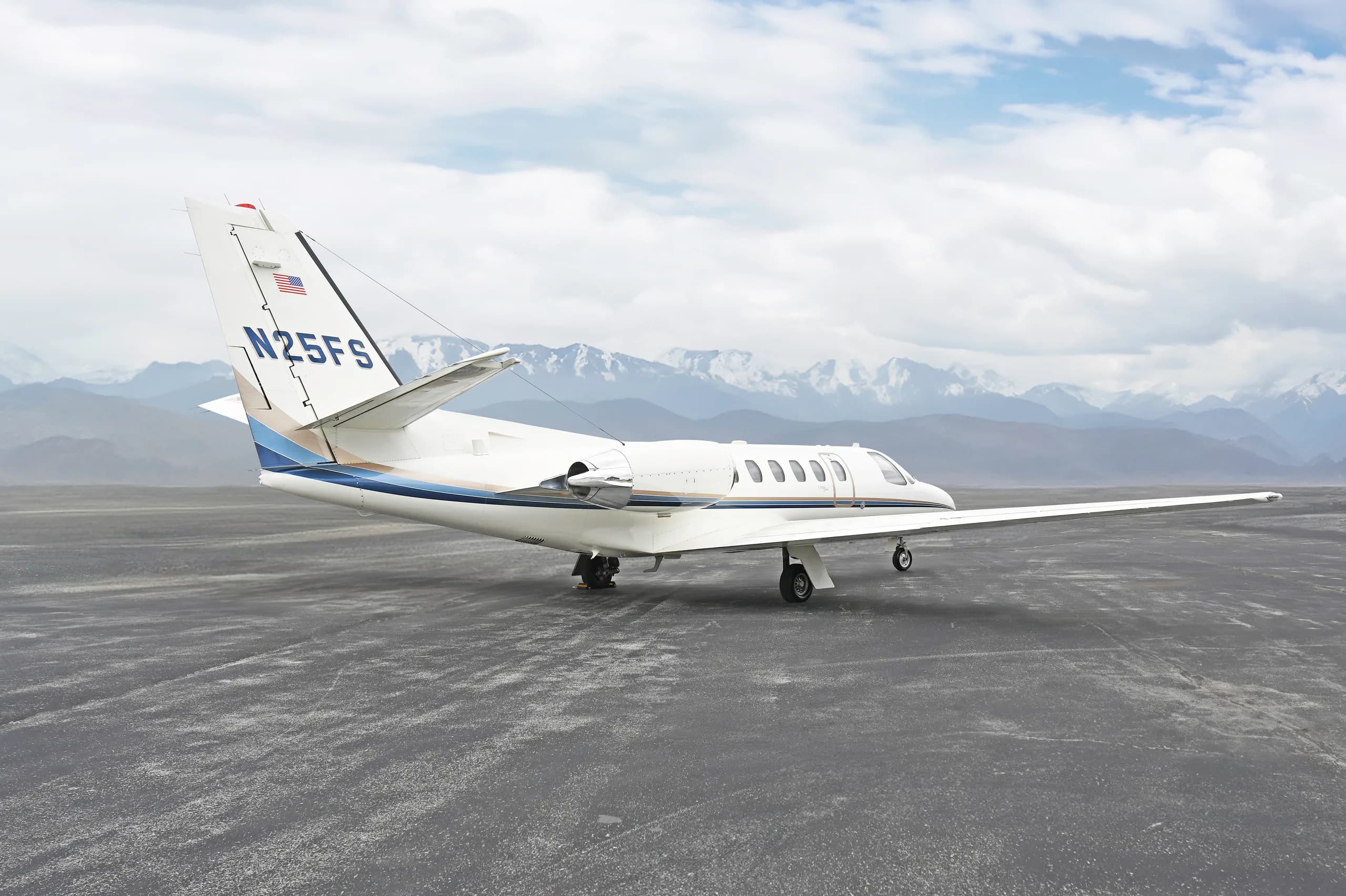 A rear three-quarter view of a white Cessna Citation business jet (N25FS) with blue and gold stripes, parked on an asphalt surface with snow-capped mountains in the background.