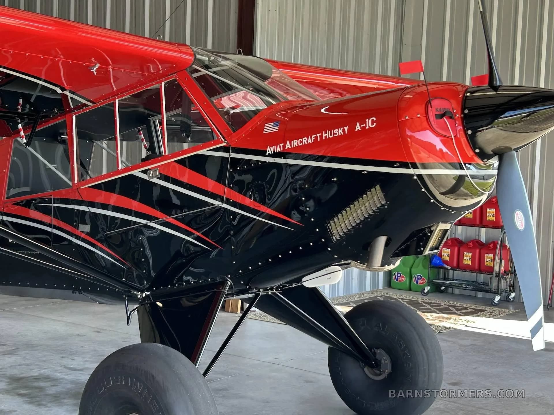 A close-up view of the red and black Aviat Husky A-1C's nose and propeller, showing the engine cowling and large tires inside a hangar.