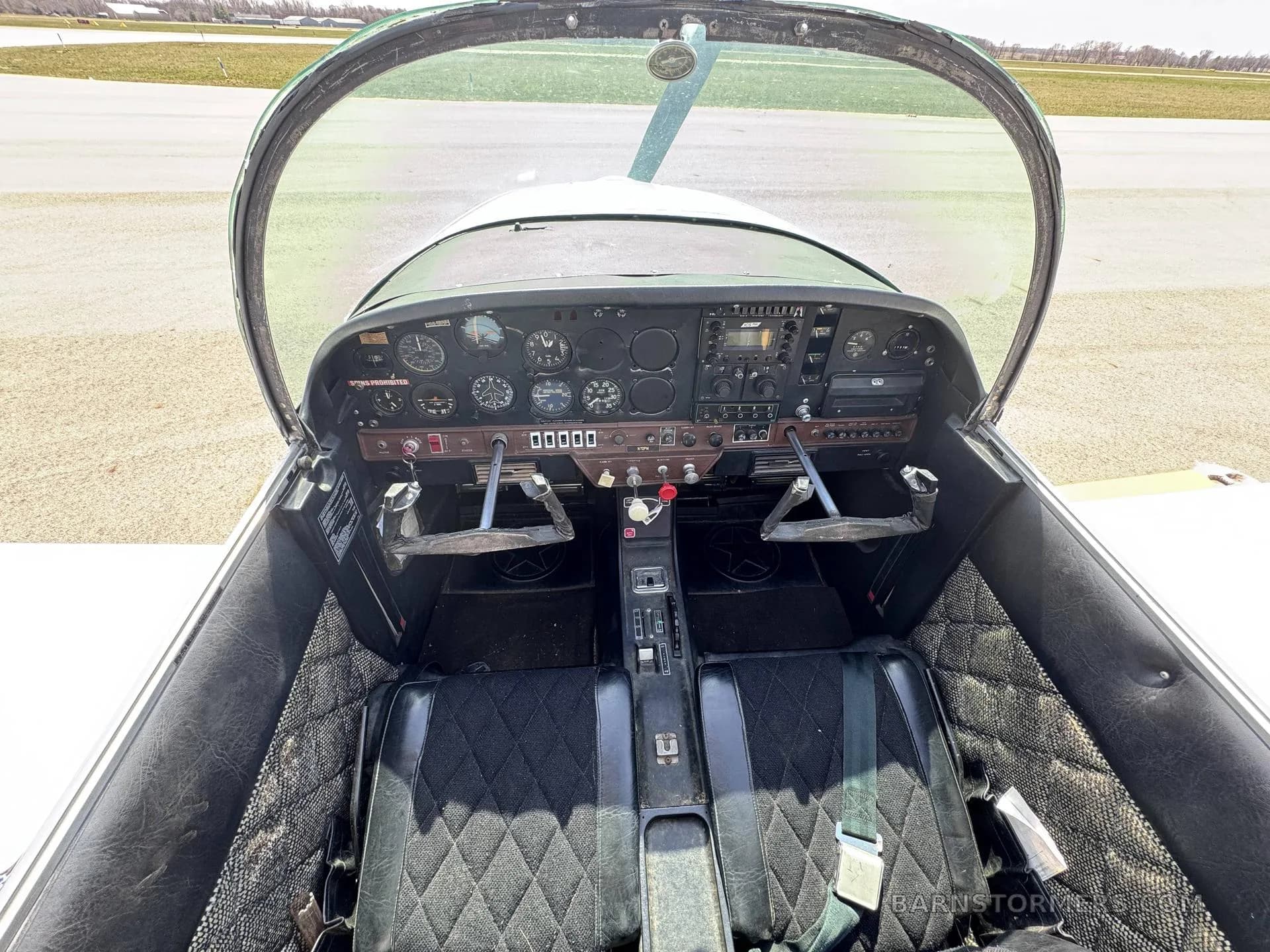 The cockpit of a Grumman American AA-5 Traveler aircraft, showing the instrument panel, yokes, and two black quilted seats.