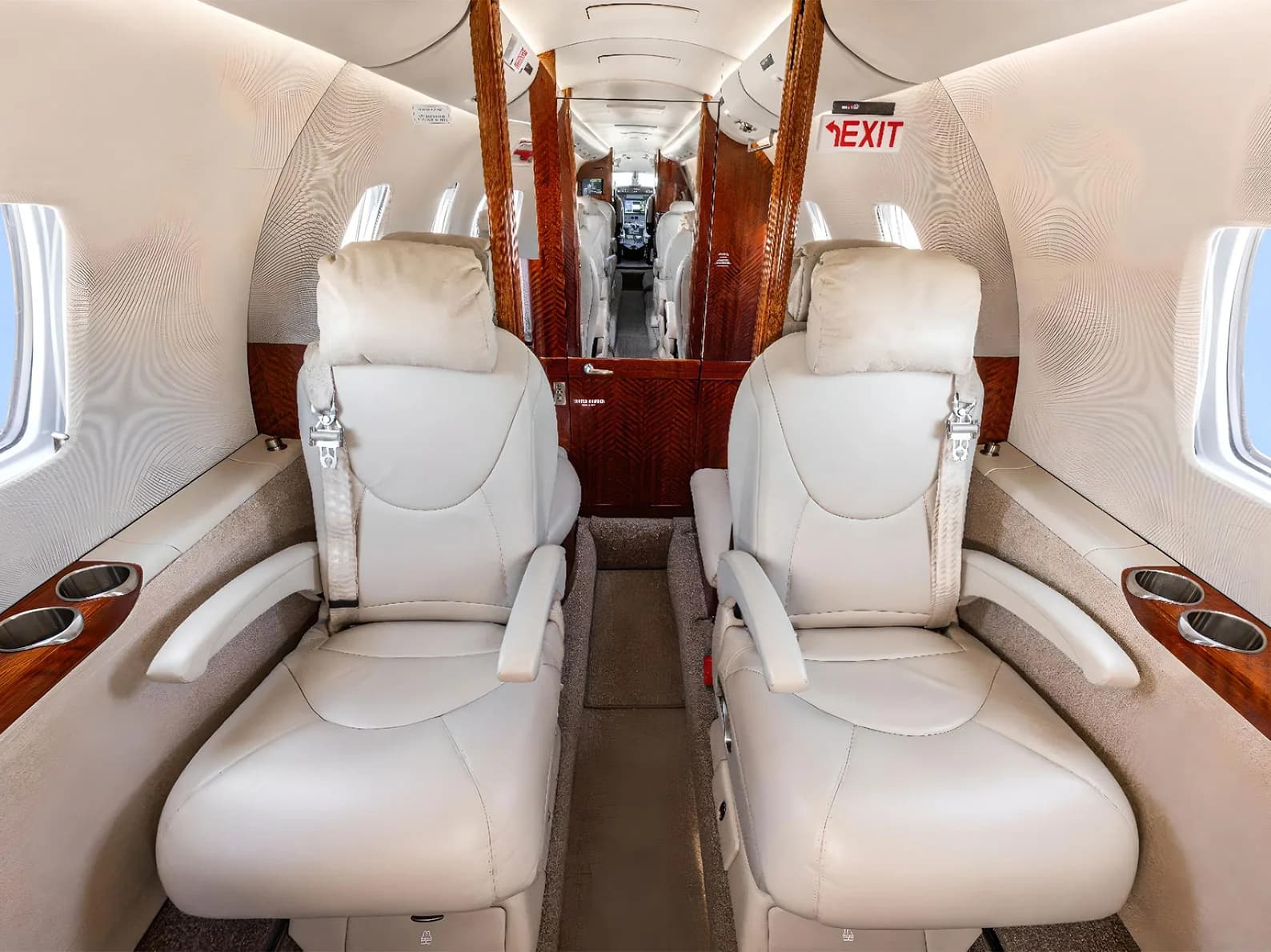 A view down the aisle of a private jet cabin, showing two forward-facing light leather seats and wood paneling.