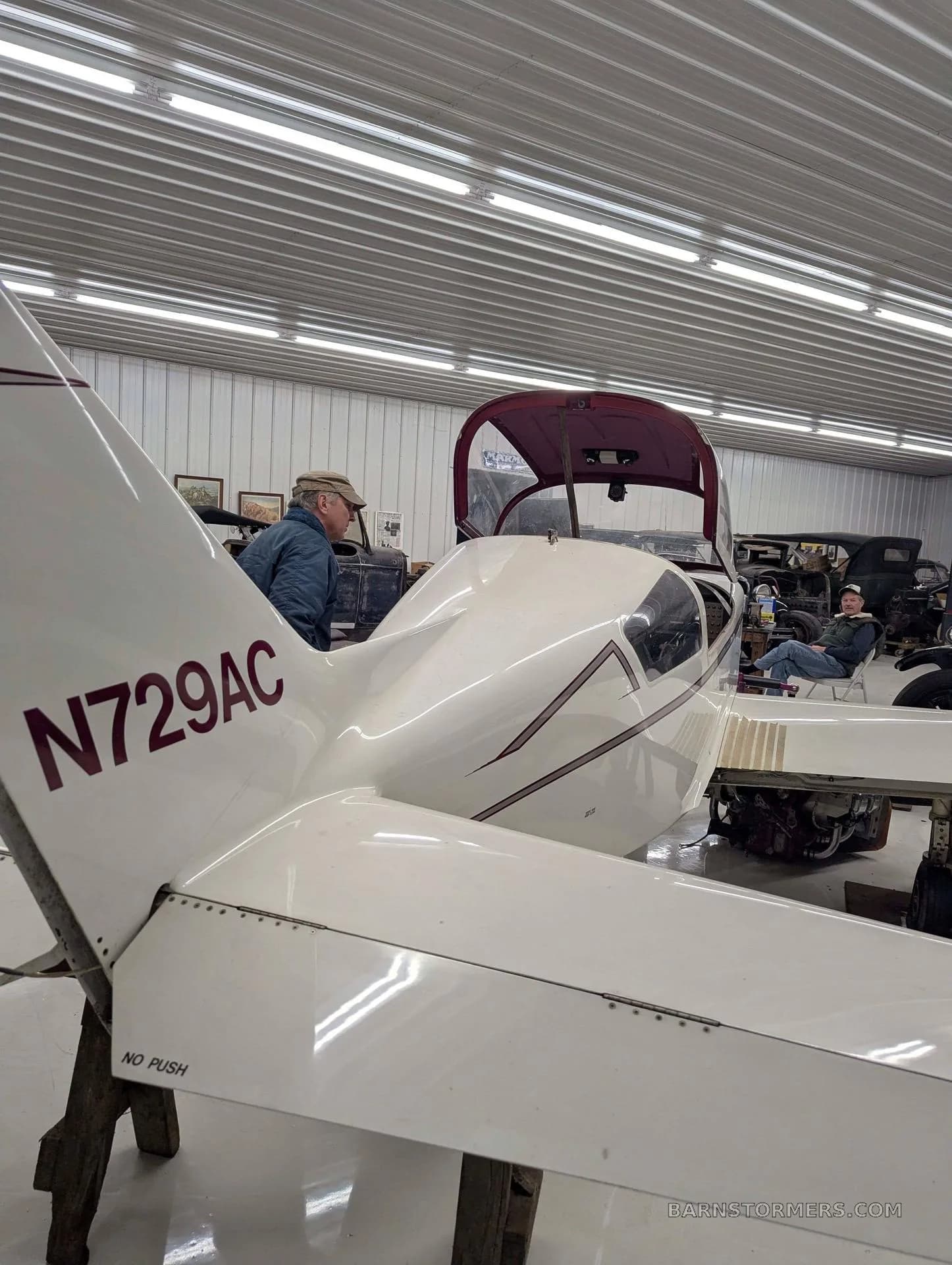 The tail section of a white aircraft with registration N729AC visible, parked in a hangar with two people.