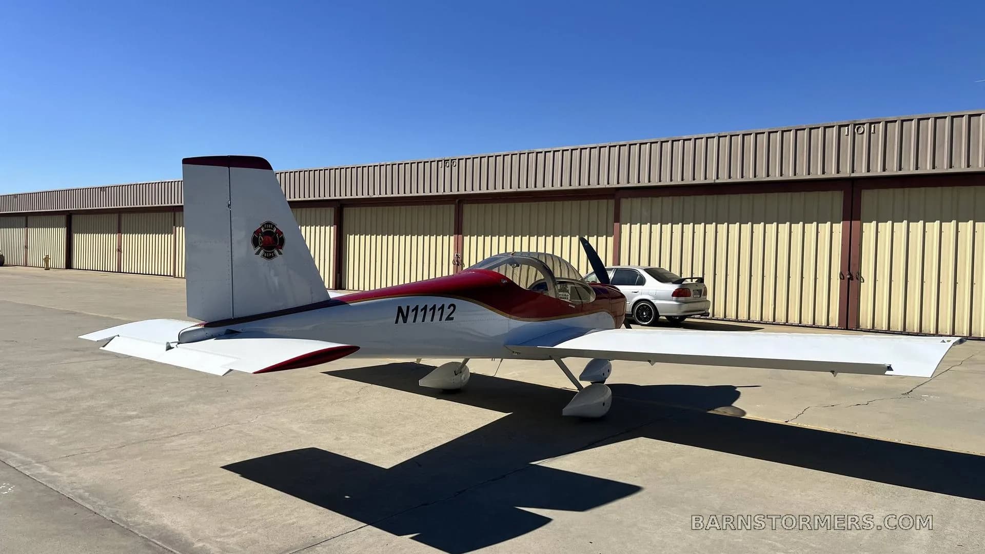 A red and white single-engine aircraft with registration N11112 is parked on an airfield apron, viewed from the rear-right with a car in the background.