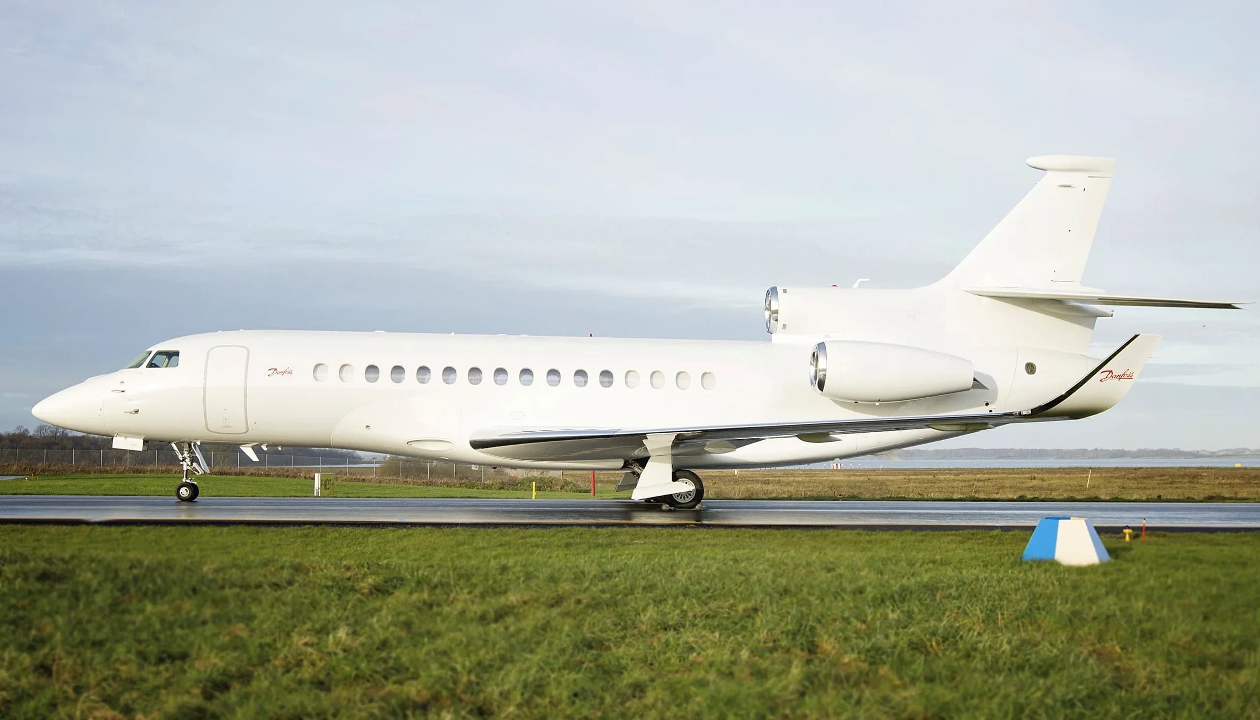 A white Dassault Falcon business jet with 'Danfoss' branding is parked on a tarmac with green grass in the foreground.